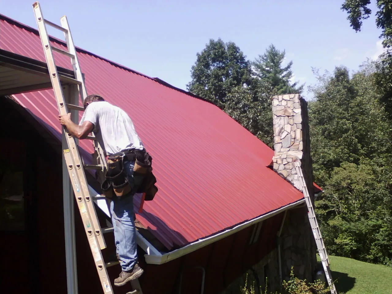 Roofer on a ladder beside a red metal roof and stone chimney — Soffit & Fascia Repair by Roofing Brothers in Lamar